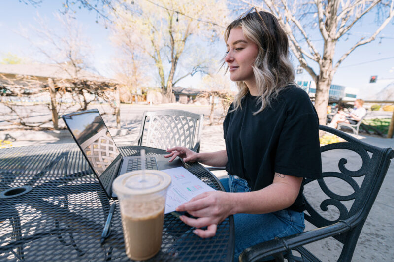 Wide Angle View of a Young Woman Telecommuting using a Laptop Outdoors on an Apartment or Cafe Patio in Spring